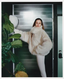 Model in fluffy cream coat posing beside large green leaves against a slatted door for Revolve by Agata Serge