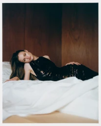 Model in a black sequin dress reclining on a white bed with dark wood paneling in the background