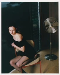 A woman in a black off-the-shoulder dress seated backwards on a wooden chair next to a standing metal fan.