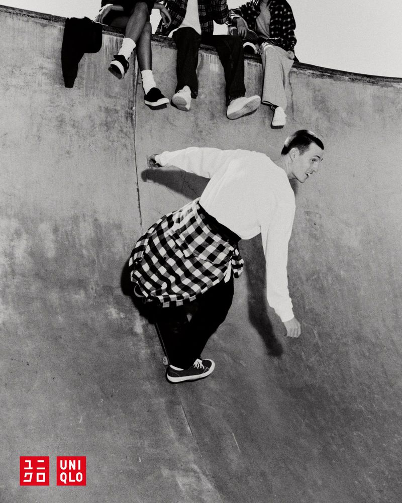 Monochromatic shot of a skateboarder riding inside a skatepark's bowl with friends sitting along the edge, wearing a flannel shirt.
