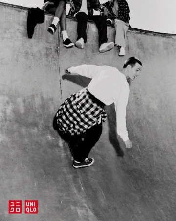 Monochromatic shot of a skateboarder riding inside a skatepark's bowl with friends sitting along the edge, wearing a flannel shirt.