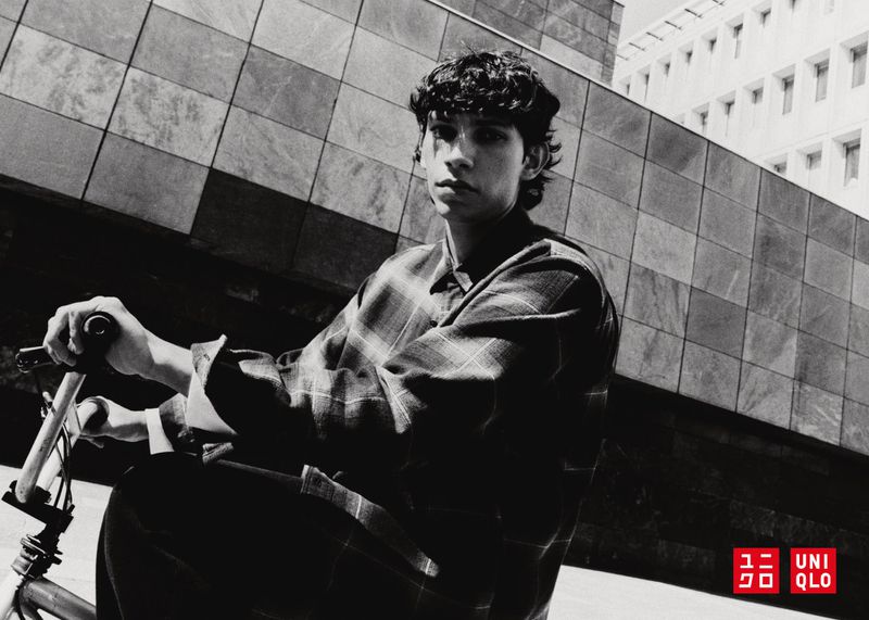 Monochrome shot of a young man with curly hair riding a bicycle outdoors against a modern building with geometric patterns and logo detail.