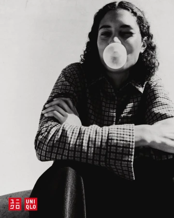 Monochrome portrait of a young adult woman with curly hair blowing a bubblegum bubble, wearing a patterned shirt against a plain backdrop.