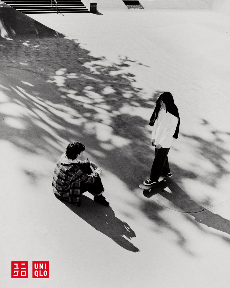 Monochrome shot of two teenagers in a skatepark, one seated while the other stands on a skateboard, casting long shadows.