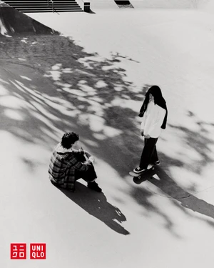 Two people at a skatepark with one person seated and another standing on a skateboard under dappled tree shadows