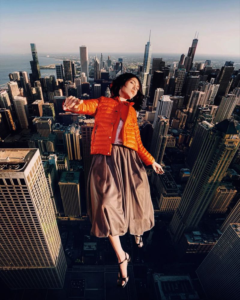 Woman in orange jacket floats above a sprawling cityscape with tall buildings and blue water, creating a surreal and captivating image.