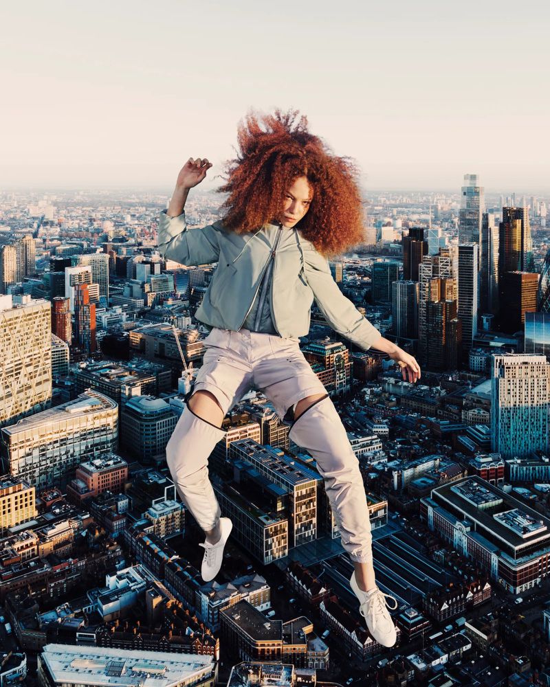 A young red-haired woman models in fashionable clothing, poised in mid-air above a cityscape in this dynamic outdoor fashion editorial.