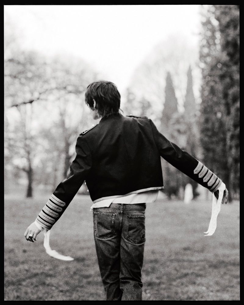 Back view of a person with dark hair wearing a jacket and jeans, holding white fabric in a park-like setting with trees in the background.