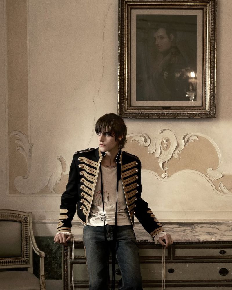 Portrait of a young adult with a black and gold military-style jacket, standing indoors with a painting on the wall.