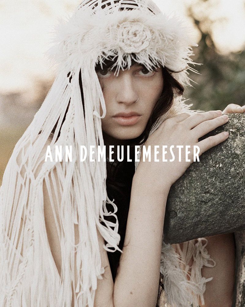 A woman with dark hair wears an elaborate white woven headpiece and fringed shawl, posing with her hand on a weathered stone in daylight.