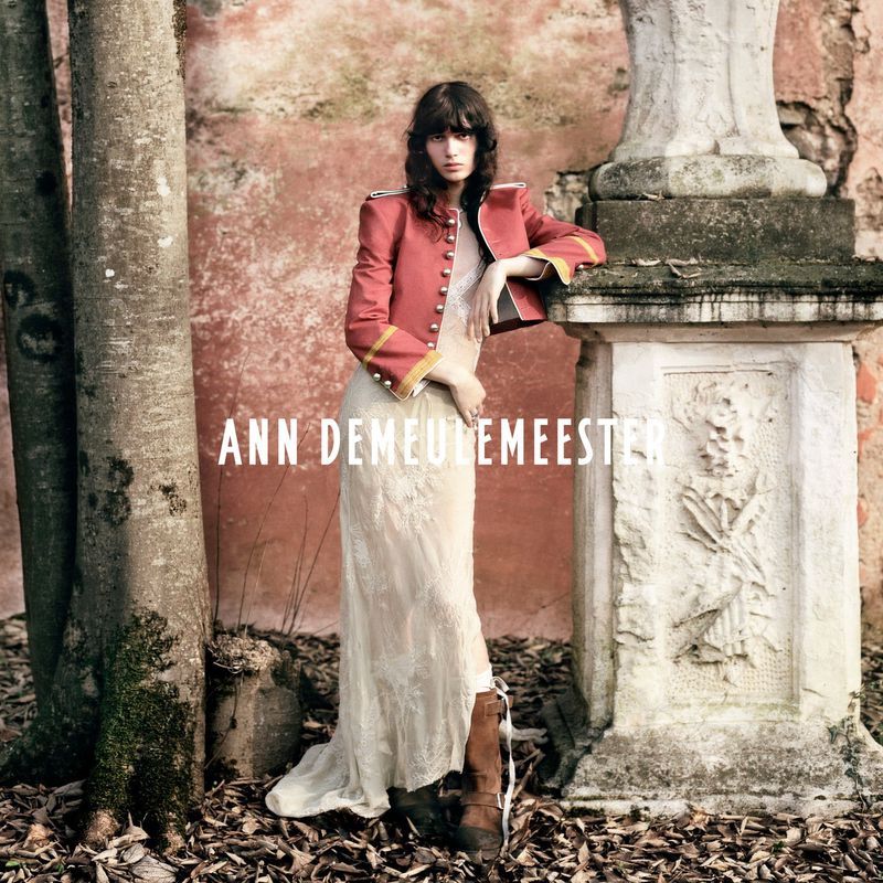 An adult female fashion model poses outdoors in a red jacket, dress, and boots, next to a decorative stone wall, with foliage visible.