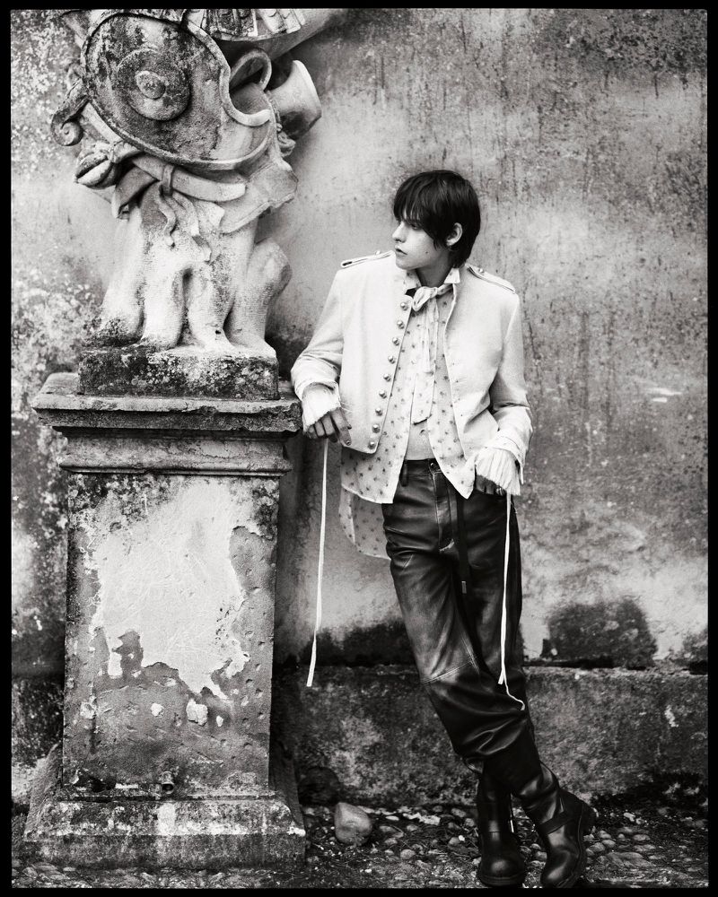 Monochrome portrait of a young adult in vintage-style clothing leaning against an ornate stone column outdoors in soft, diffused light.
