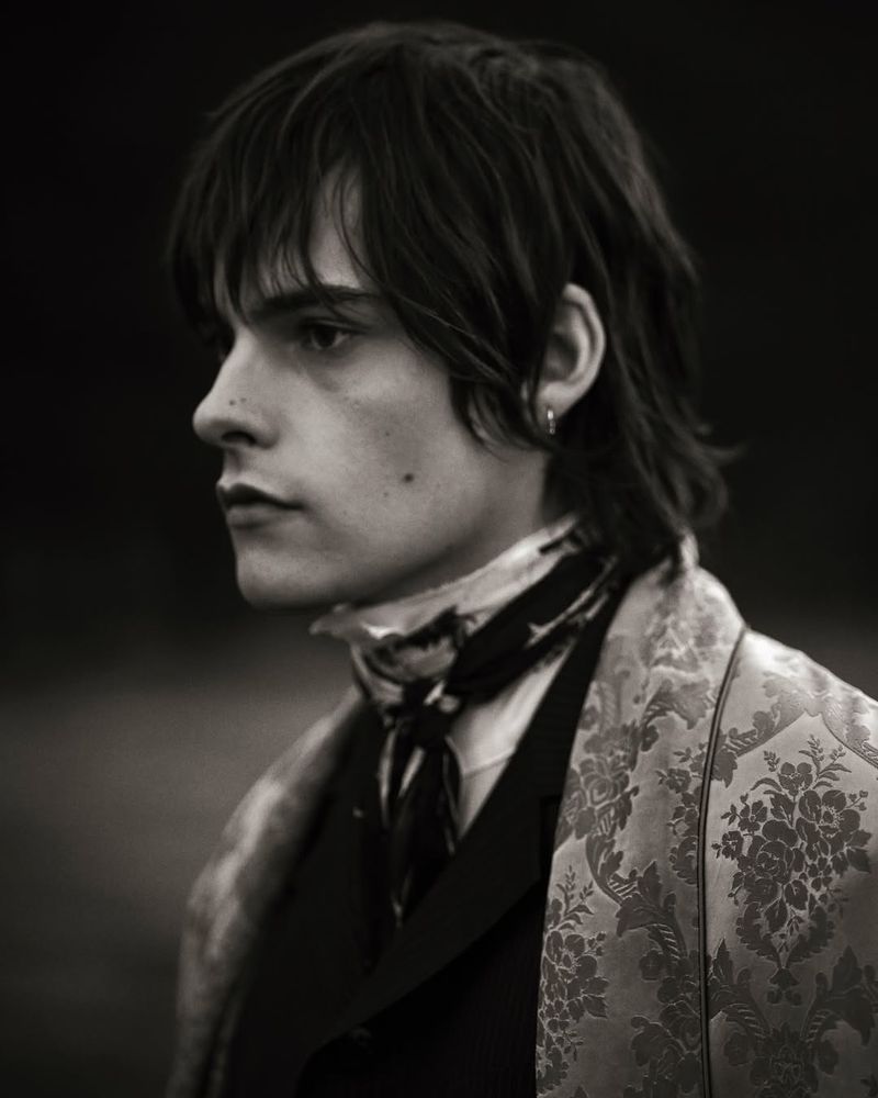 Close-up profile of a man with tousled hair, wearing a patterned floral shawl over a dark coat and a dark neck scarf.