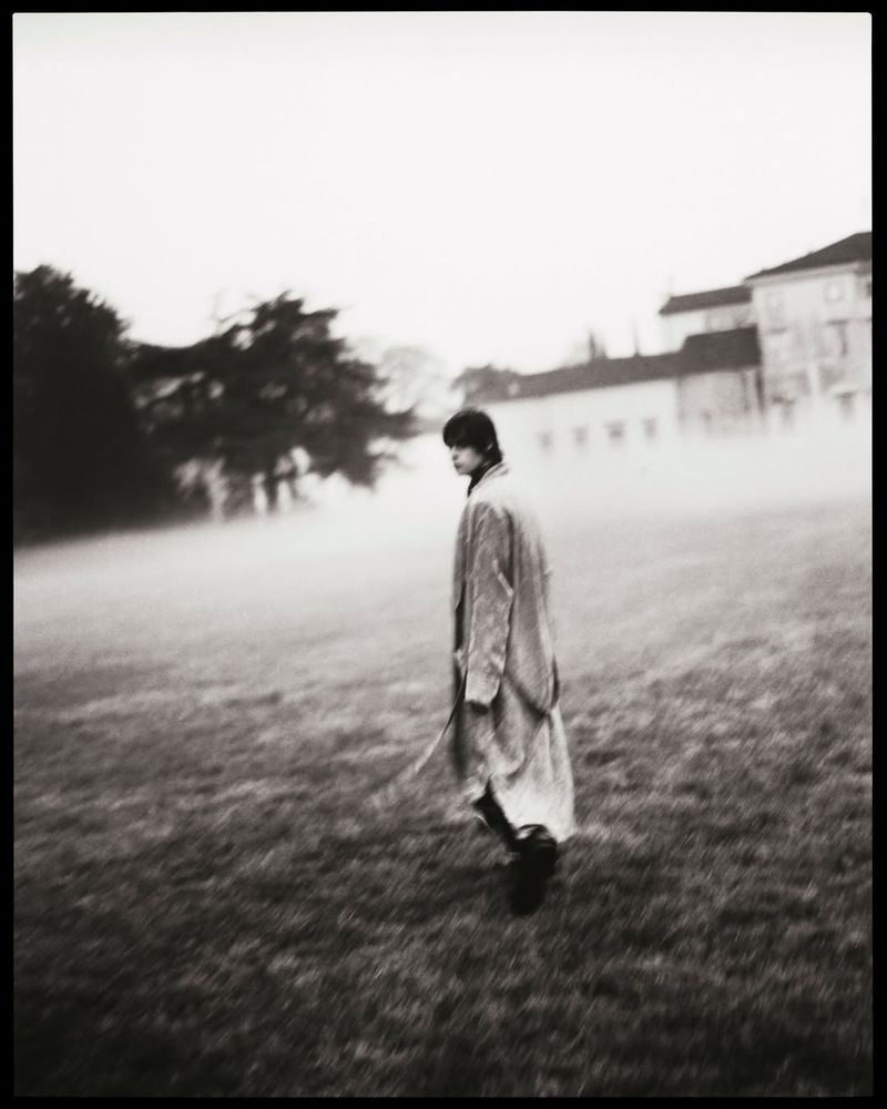 Woman walks through a grassy field in a long coat on an overcast day. Trees and a building are partially visible in the background.