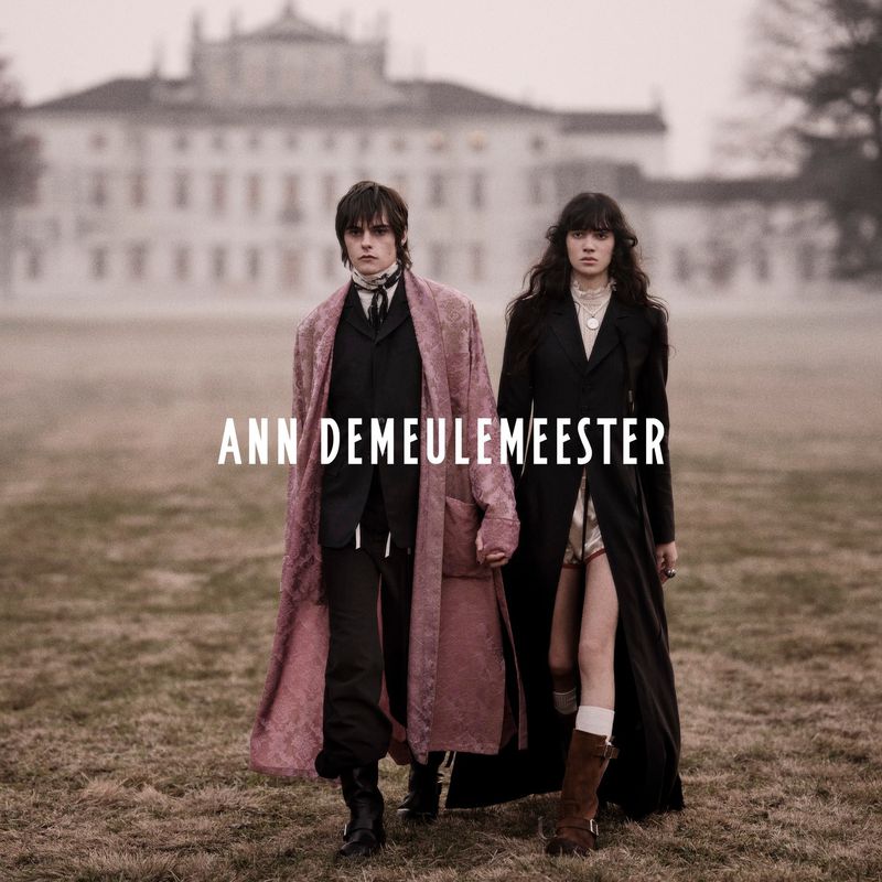 A fashion editorial photograph features two models in elegant coats walking on a grassy field with a grand building in the background.