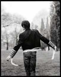 Monochrome fashion campaign image of a model from behind wearing a dark military-style jacket with white sleeve ribbons in a park.