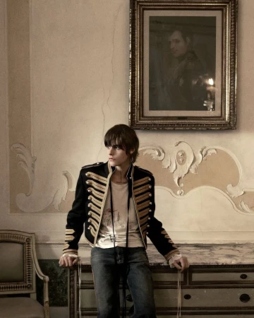 Model in a black military-style jacket with gold braiding leaning against an ornate credenza in a classical interior.