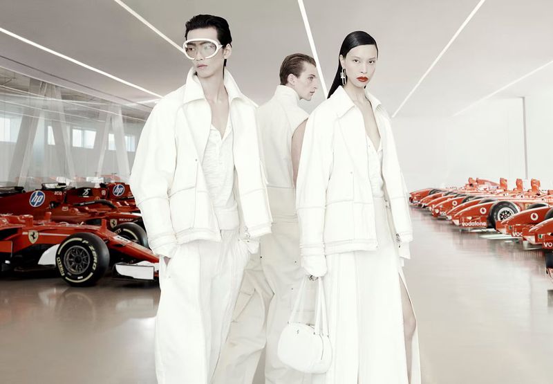 Two fashion models pose in white outfits alongside red Ferrari race cars in a bright, modern showroom with light flooring and walls.