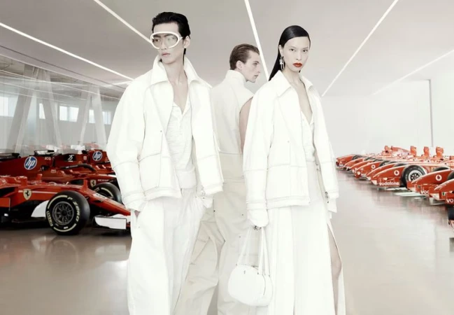 Two fashion models pose in white outfits alongside red Ferrari race cars in a bright, modern showroom with light flooring and walls.