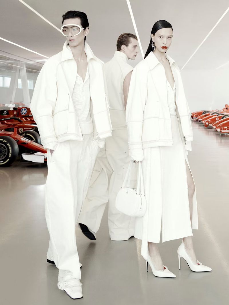 Fashion models in white clothing pose next to Ferrari race cars in a bright, minimalist setting, showcasing modern style and automotive design.