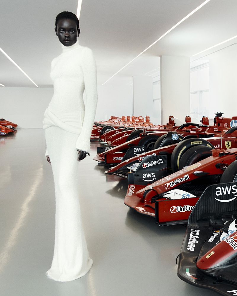 Elegant Black woman in a white dress poses in a showroom, surrounded by several red Formula One race cars, under bright white lighting.