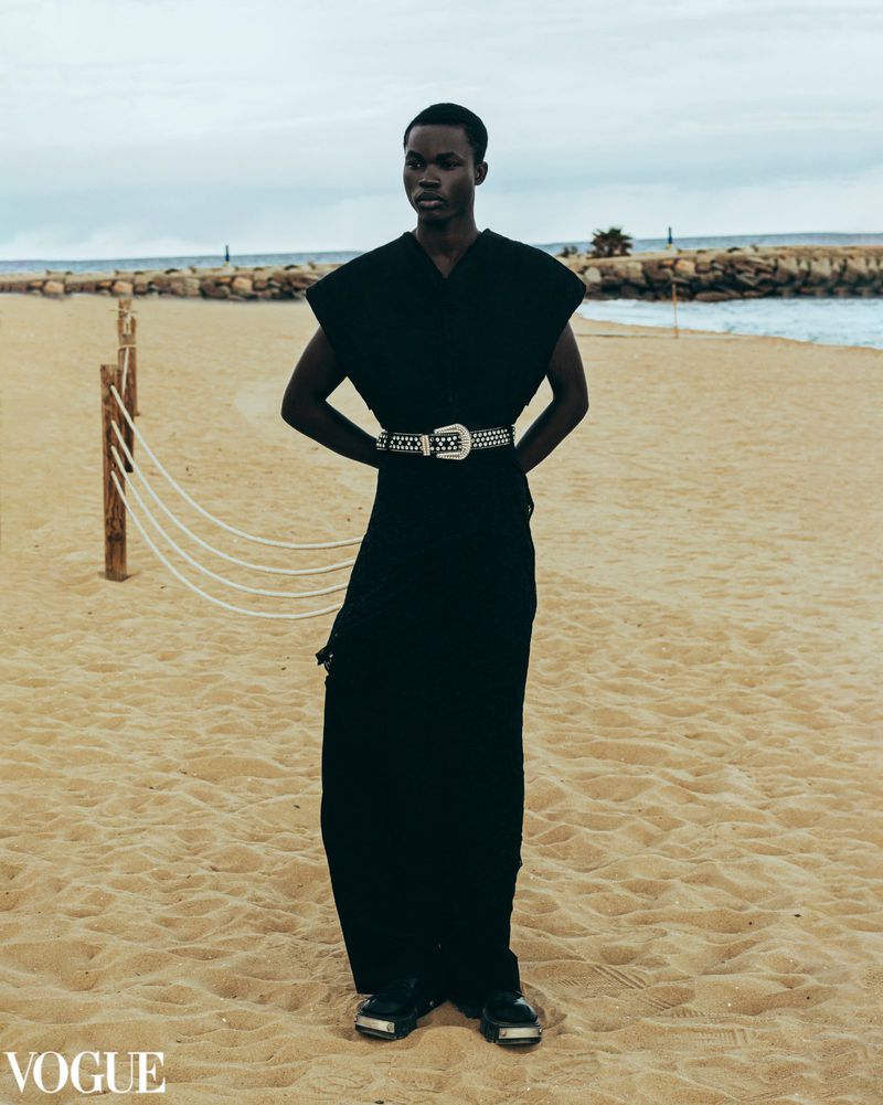 Fashion shot of a Black male model wearing a dark outfit with silver belt standing on a sandy beach with ocean and cloudy sky.