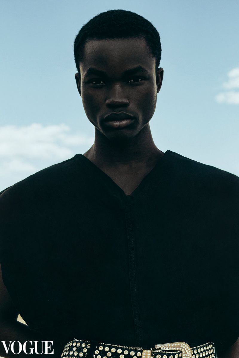 Dark-skinned male model gazes directly at the camera, wearing a black top and a studded belt against a bright blue sky background.