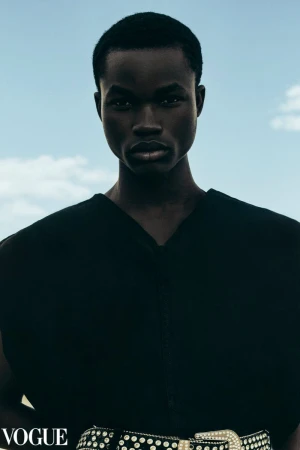 Dark-skinned male model gazes directly at the camera, wearing a black top and a studded belt against a bright blue sky background.