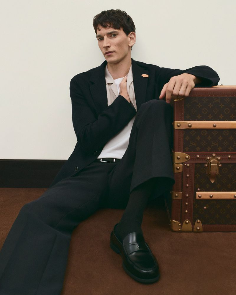 Male model poses in a black suit, white shirt, black socks and loafers, sitting by a Louis Vuitton trunk against a beige wall.