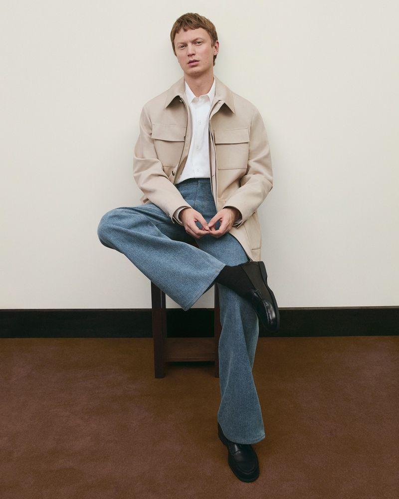 A fair-skinned man with blond hair sits on a stool, wearing jeans, a jacket, and black boots against a neutral backdrop indoors.