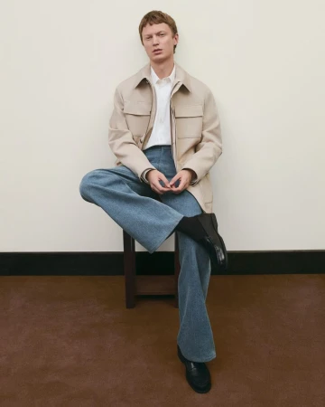 A fair-skinned man with blond hair sits on a stool, wearing jeans, a jacket, and black boots against a neutral backdrop indoors.