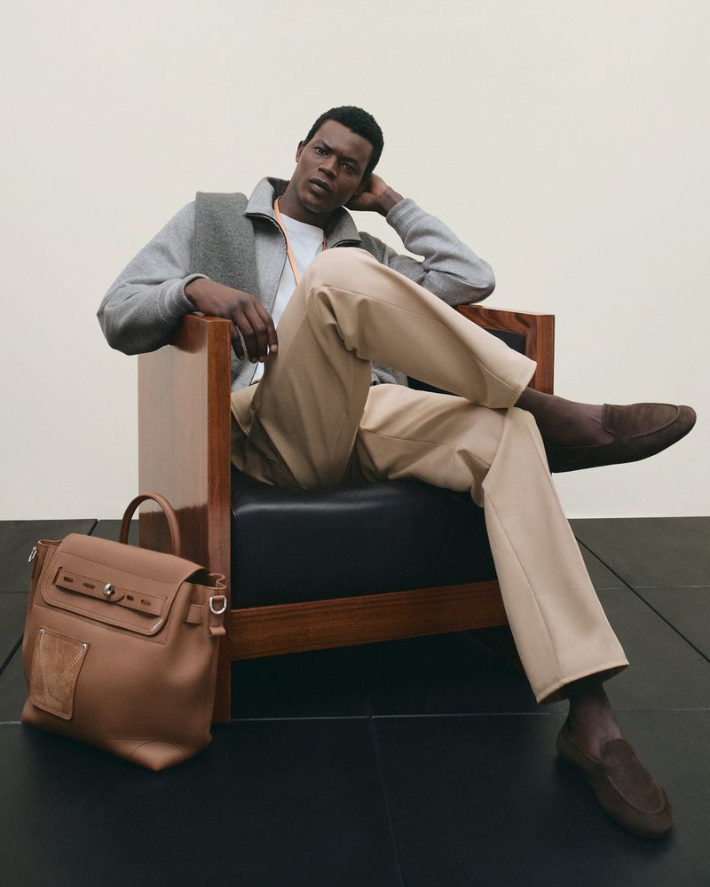 A stylish dark-skinned male model in a chair, wearing a gray jacket and tan pants, complemented by a leather satchel on the floor.