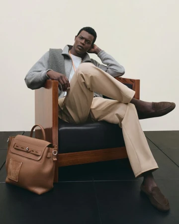 A stylish dark-skinned male model in a chair, wearing a gray jacket and tan pants, complemented by a leather satchel on the floor.