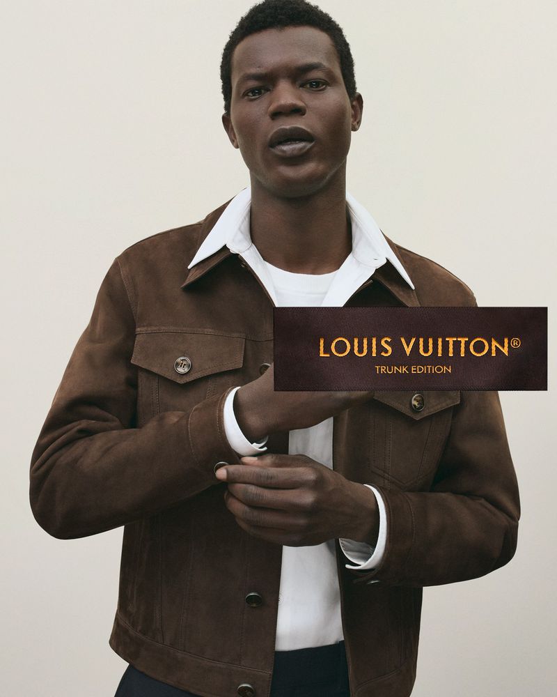 A black man in a brown jacket stands indoors, adjusting his cufflink. Louis Vuitton logo displayed. Fashion, style, and luxury are visible.