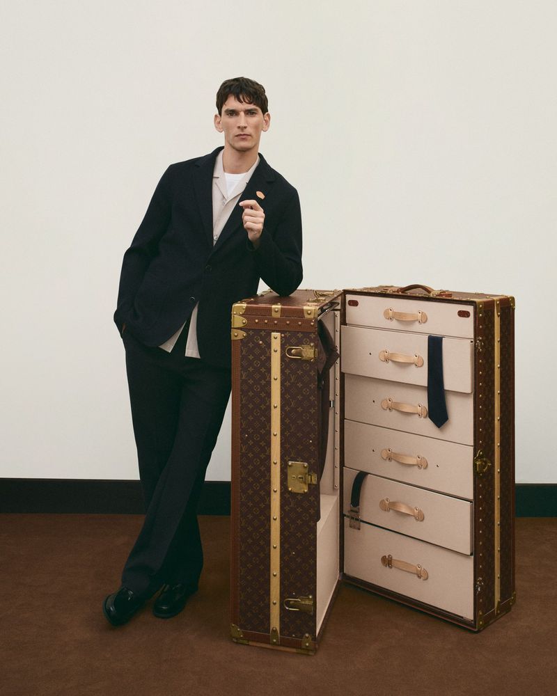 Man poses beside a brown Louis Vuitton wardrobe trunk. He wears a dark suit. The trunk is open, revealing drawers and garment storage.