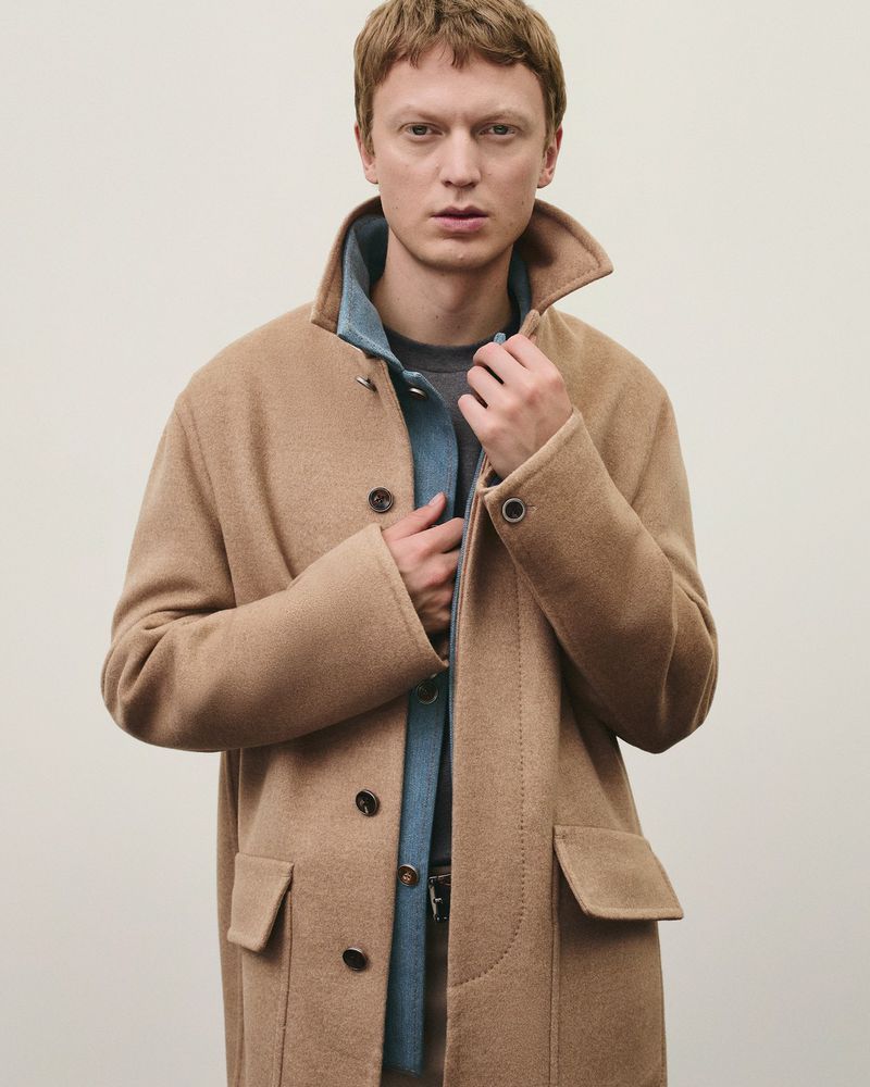 A fair-skinned adult man with red hair wears a tan overcoat over a denim shirt and gray sweater, standing indoors with a neutral backdrop.