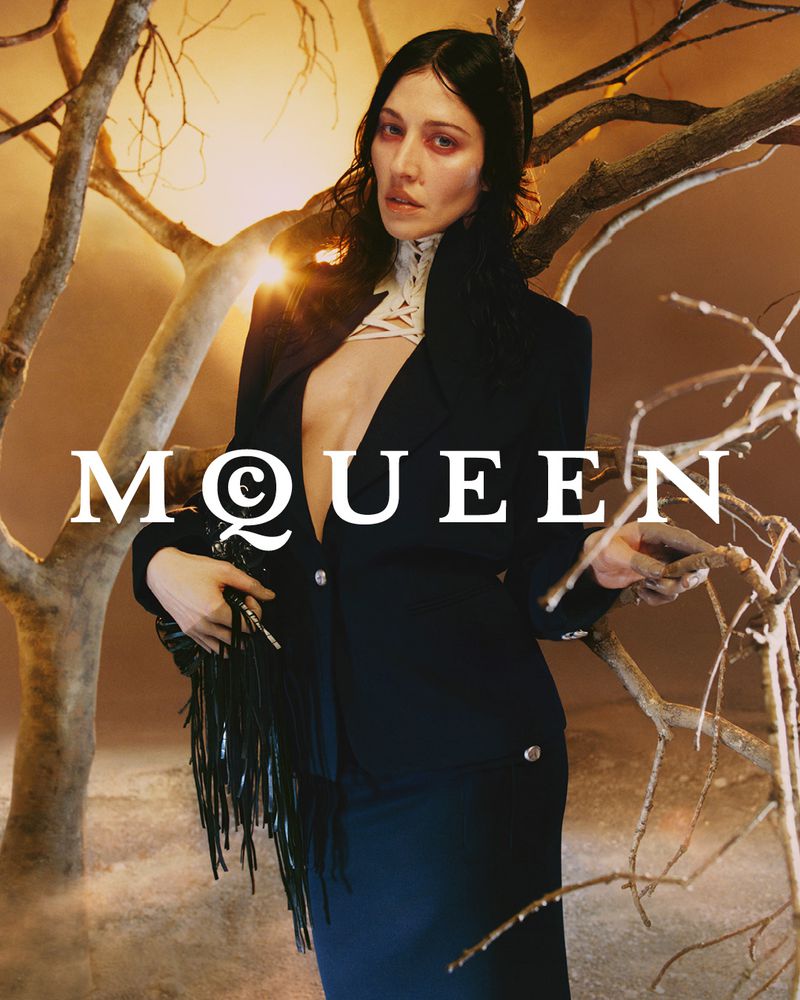 Fashion shot of a woman in a dark suit with fringed bag, posing near bare tree branches against an amber gradient background with brand text.