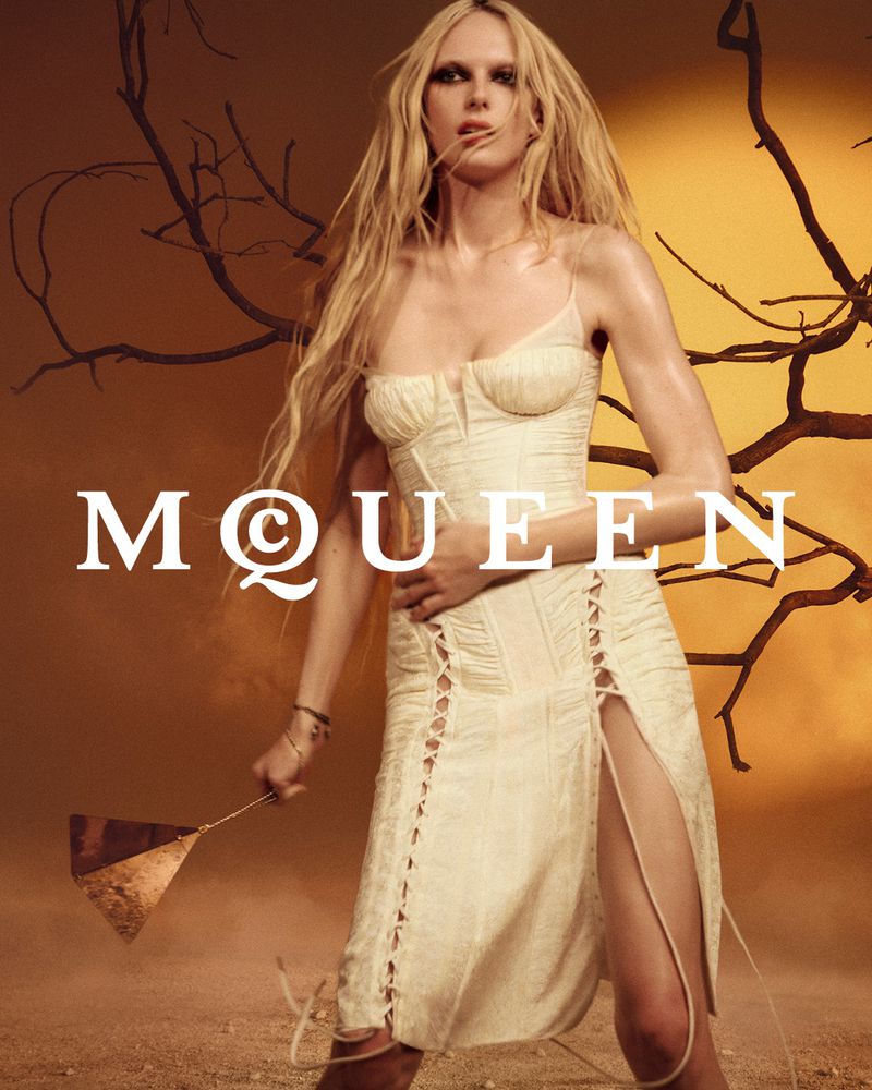Model wearing a cream corset dress with lace-up details posing outdoors against an orange sky with bare tree branches