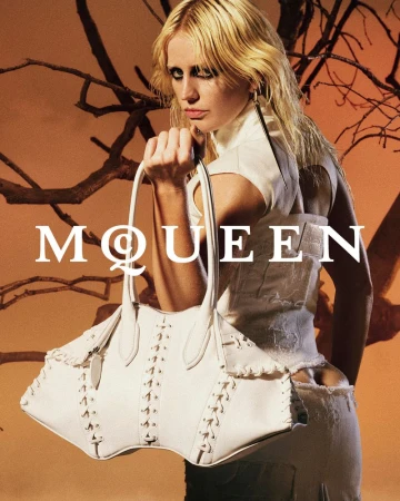 Fashion editorial shot featuring a blond woman holding a large white leather handbag in front of a blurred orange and brown background.