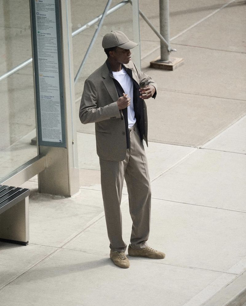 Young man in a taupe suit and cap standing at a bus stop on a city street, looking away, in a medium shot with natural lighting.