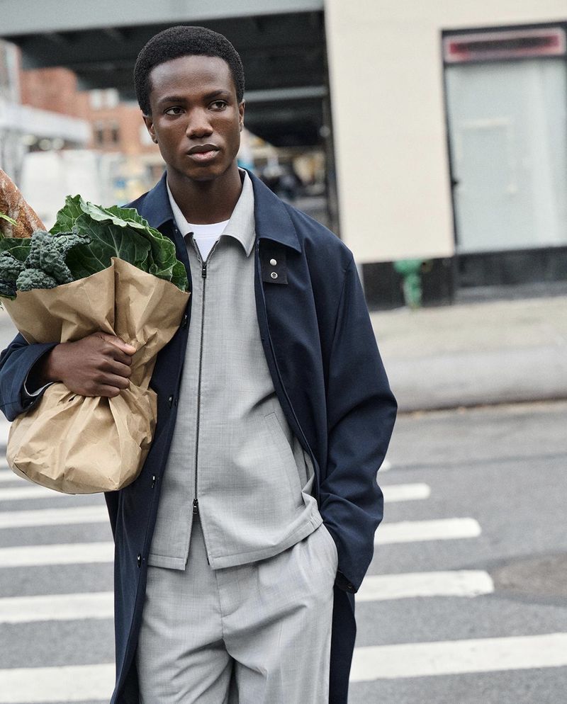 An adult man with a serious expression is holding a brown paper bag with kale and a baguette on an outdoor city street.