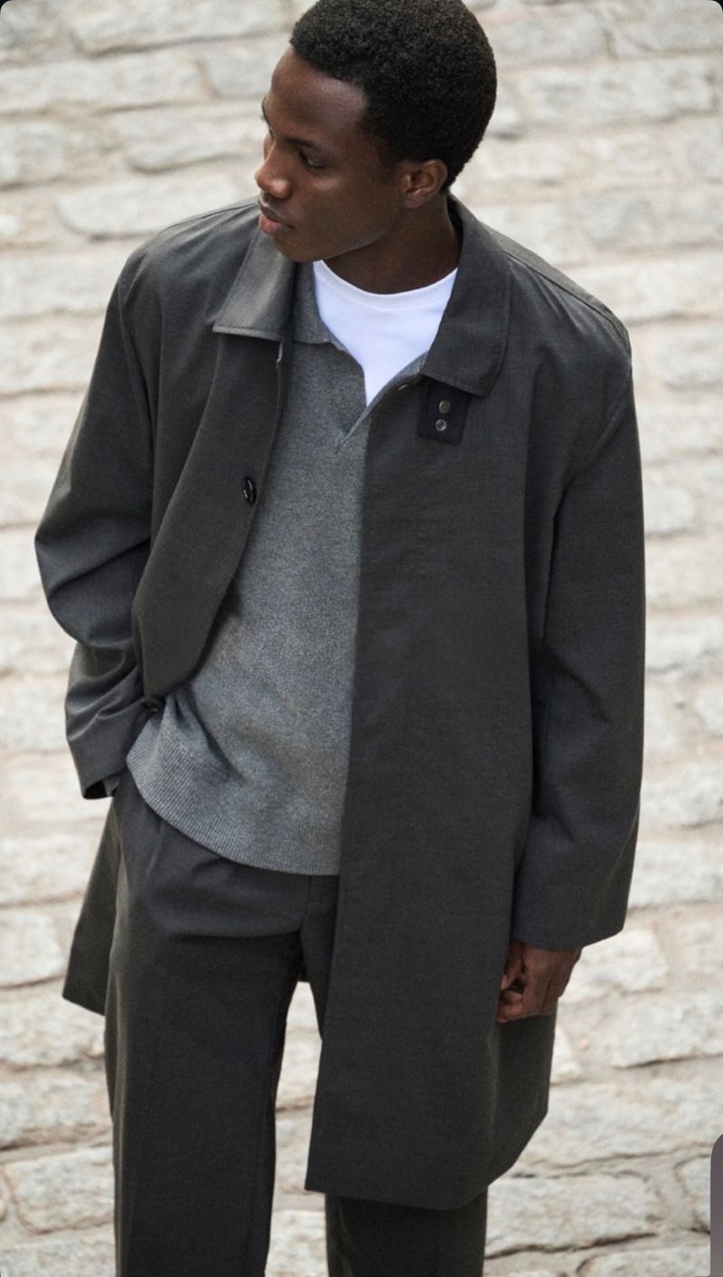 Dark-skinned male model in gray coat, sweater, and pants poses on a brick surface, looking to the left, captured in outdoor fashion shot.