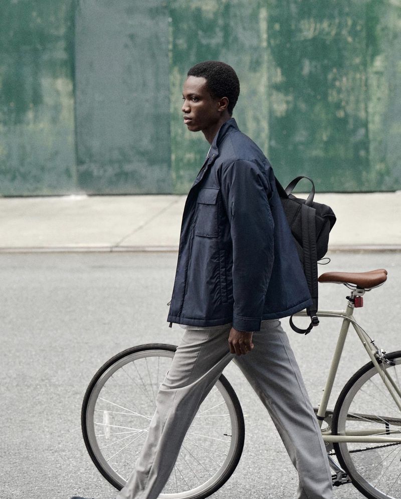 Young man in a jacket walks a bicycle on the street with an urban background. Day scene with grey and blue tones, medium shot.