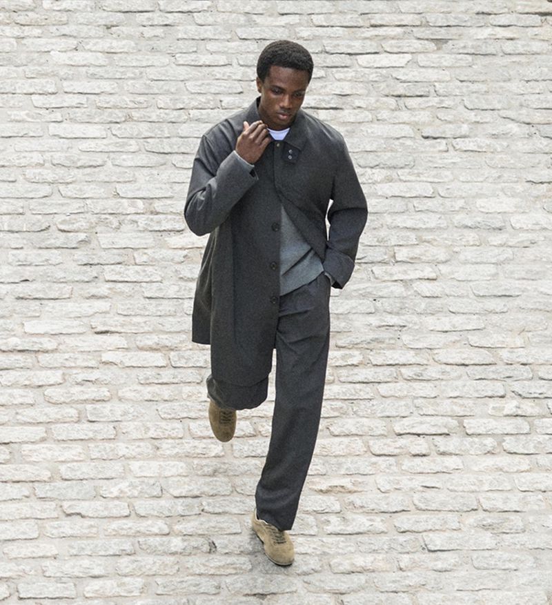 Dark-skinned man in a gray coat walks on cobblestones. He wears gray pants, a gray sweater, and tan shoes. High-angle shot, outdoor fashion.