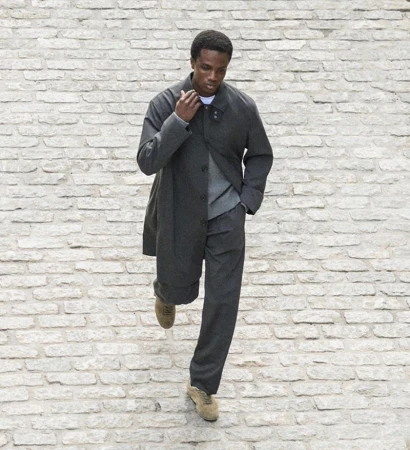 Dark-skinned man in a gray coat walks on cobblestones. He wears gray pants, a gray sweater, and tan shoes. High-angle shot, outdoor fashion.