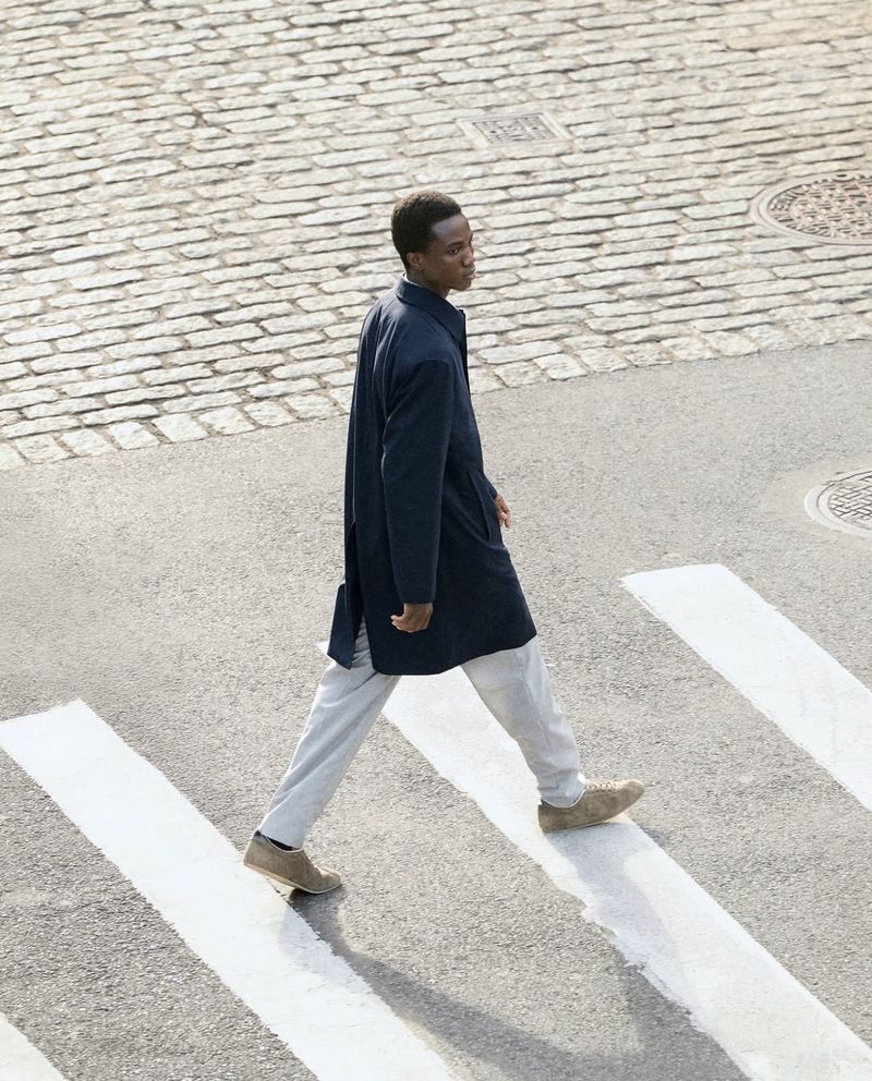 A black male adult walks across a pedestrian crossing with white stripes, wearing a dark coat and light trousers on a cobblestone street.