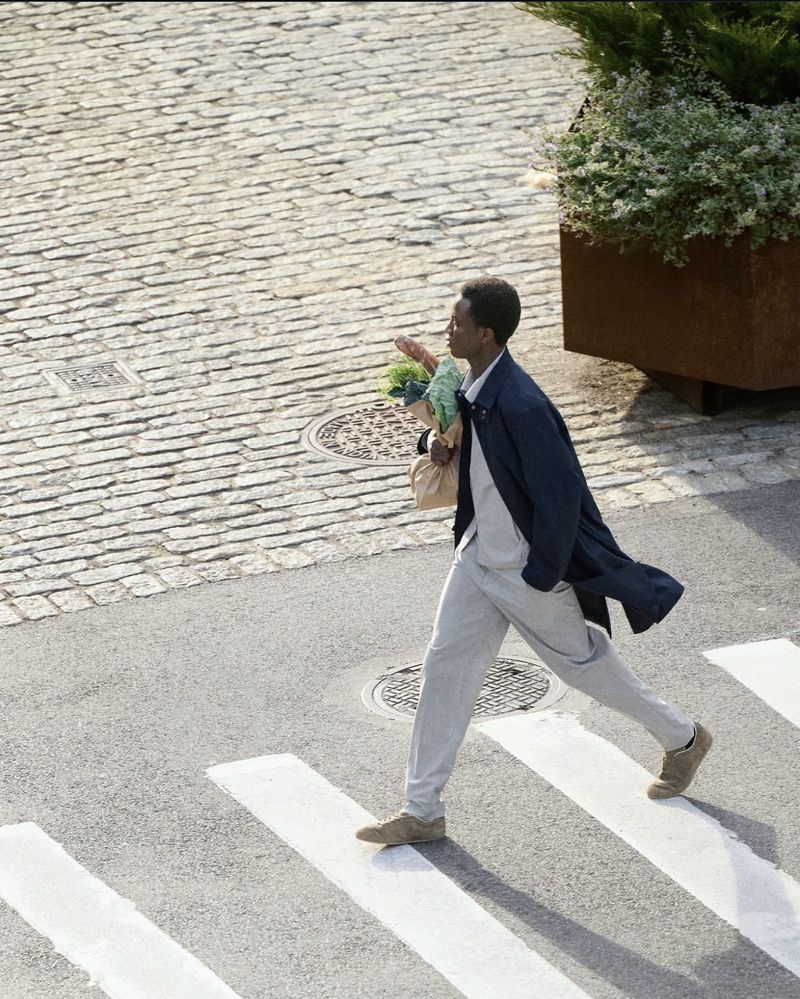 An African American man in a trench coat walks across a crosswalk with greenery and brickwork in the background on a sunny day.