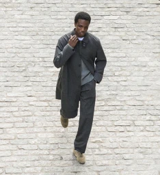 Man wearing a dark grey coat and trousers walking against a pale textured stone wall background
