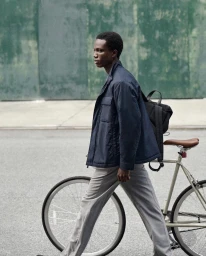 Black man walking past a green wall next to a bicycle while wearing a dark jacket and grey trousers