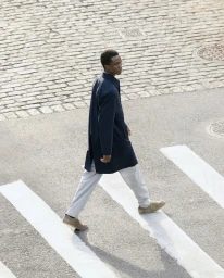 A Black man wearing a dark coat and light trousers walking across a paved street with white crosswalk lines